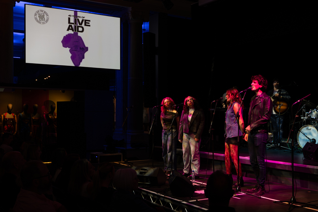 4 vocalists stand together on stage while one sings, surrounded by Live Aid imagery on the screens.