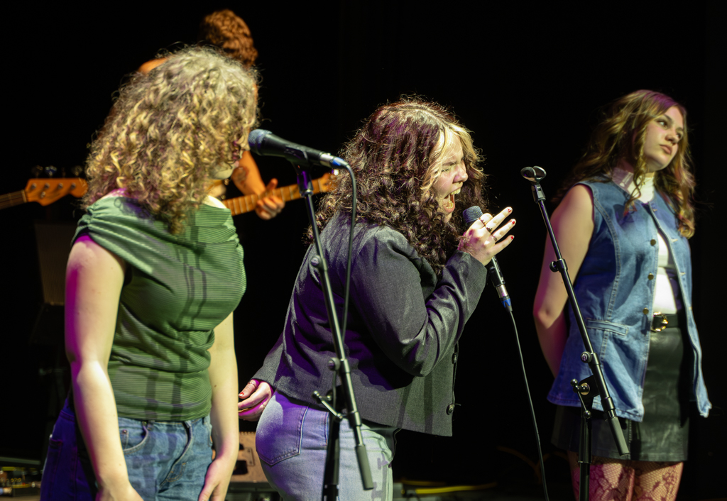 Three singers stand together as the central singer sings a solo.
