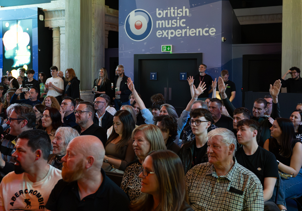 Audience in the Cunard building watching the Live Aid tribute with a British Music Experience sign at the back. Some audience members have their hands right up in the air.