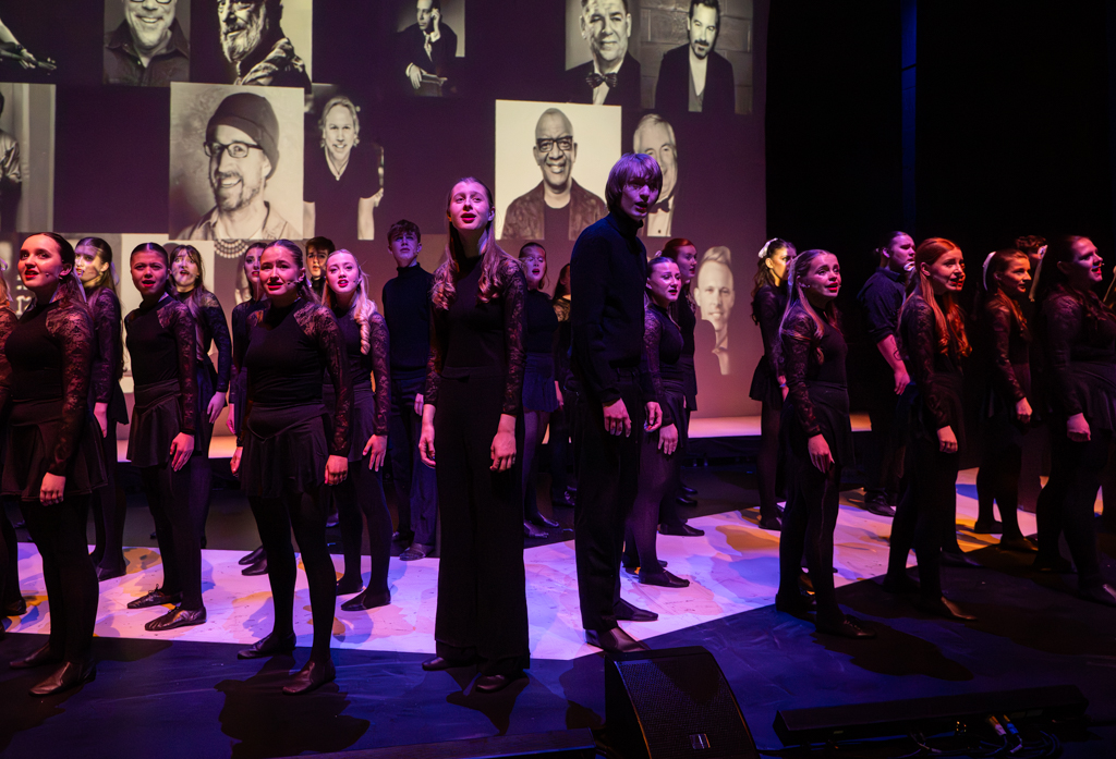 A large group of students look to the crowd, standing at an angle, whilst singing.
