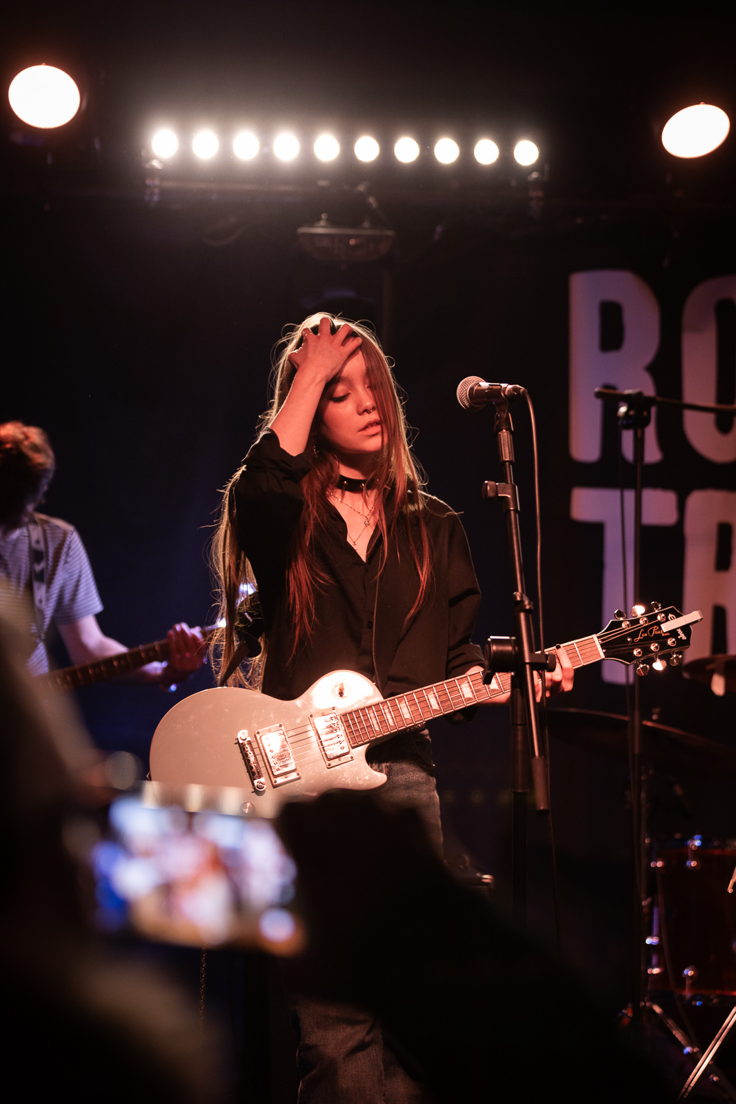 A Music student brushes their hair back whilst performing, playing guitar on stage.