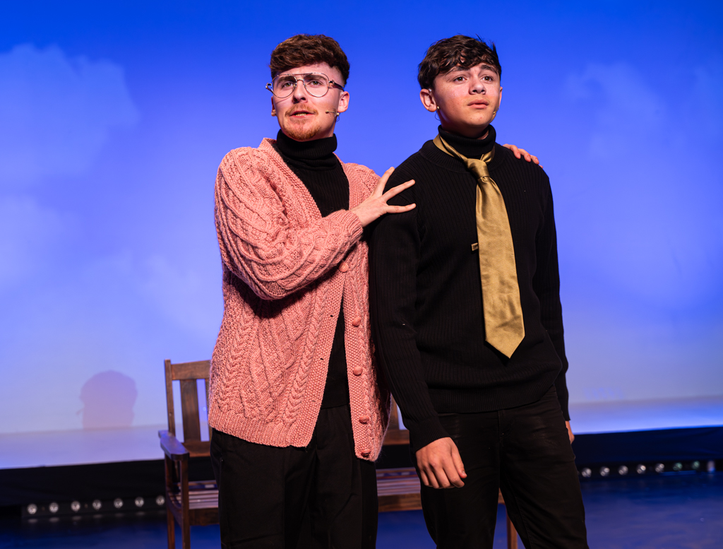 Two students stand together on stage, one with a cardigan on and glasses, the other with a tie around their neck. 
